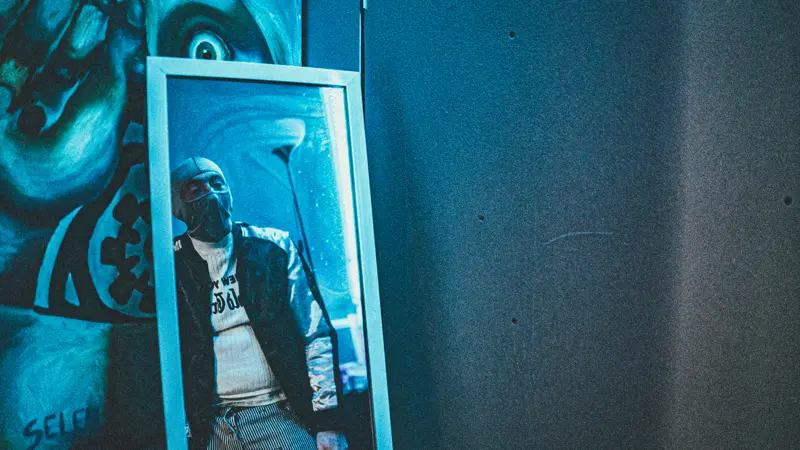 Captain Real Estate in a balaclava and leather jacket, reflected in a mirror leaning against a mural
