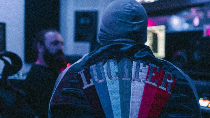 Captain Real Estate from behind in a hooded mask and a jacket embroidered with the word Lucifer, inside a studio