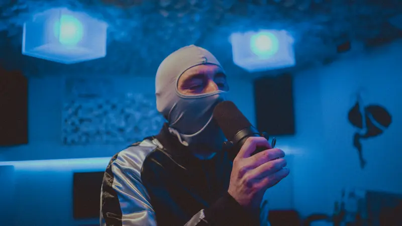 Captain Real Estate in a white balaclava rapping into a microphone under blue light